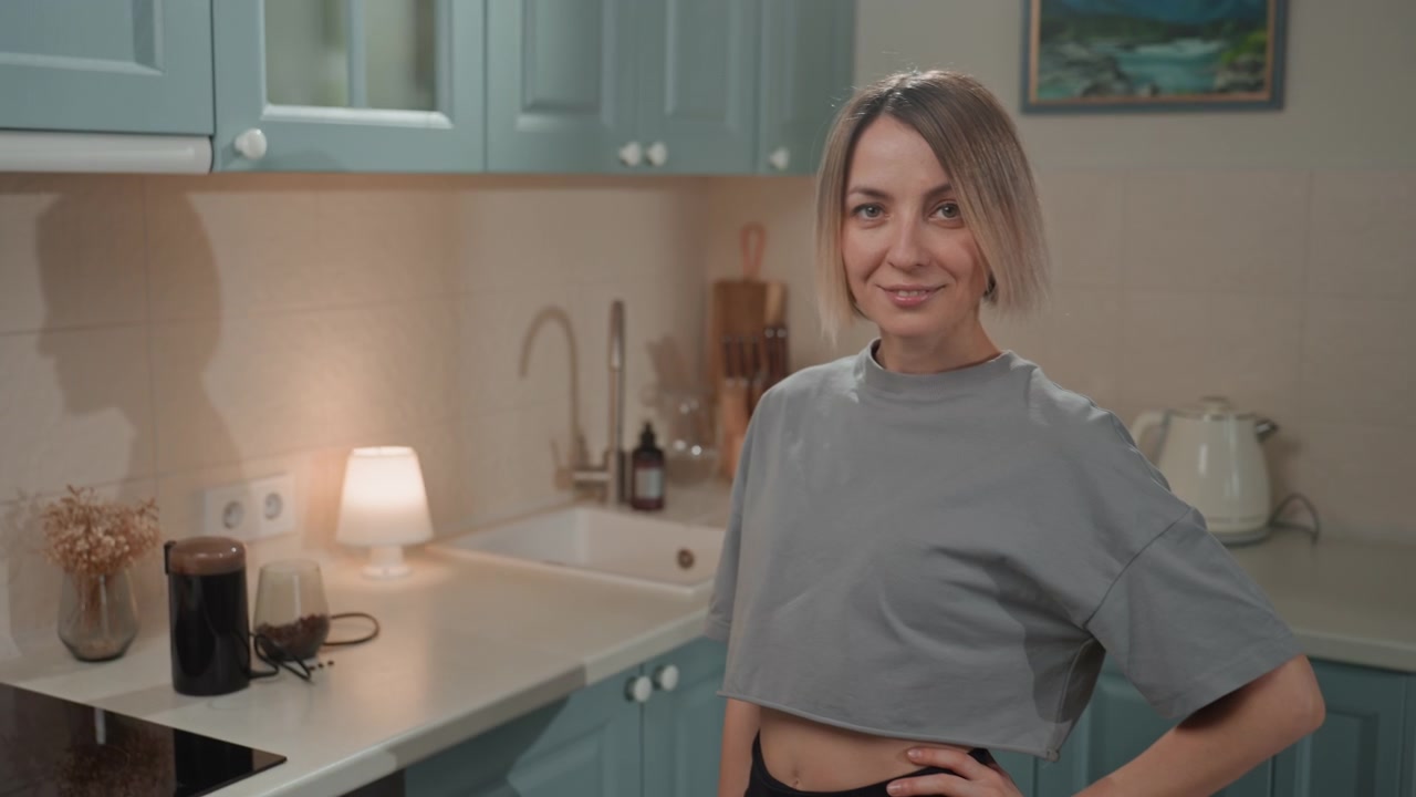 Giovane donna caucasica in cucina, che sorride e posa accanto a mobili color pastello e alla luce soffusa di una lampada, atmosfera mattutina rilassata con taglio a caschetto e maglietta corta, atmosfera intima e delicata, content creator casalinga