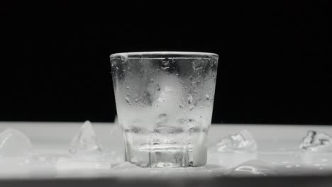 Hielo-Congelado-Cayendo-En-Un-Vaso-Mojado-Sobre-Una-Mesa-Blanca