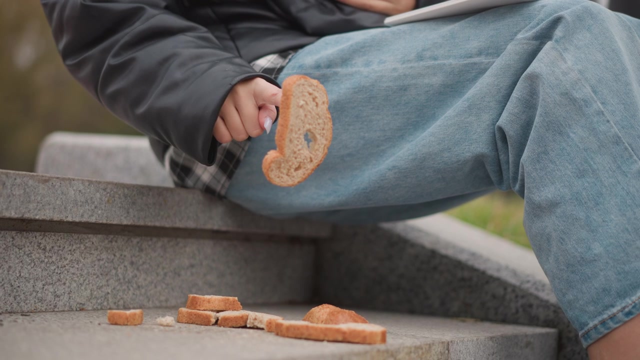 Close up of young woman sitting on stone steps picking up and examining pieces of bread. Woman wears casual clothing including blue jeans while holding food with her hand. scene is outdoors