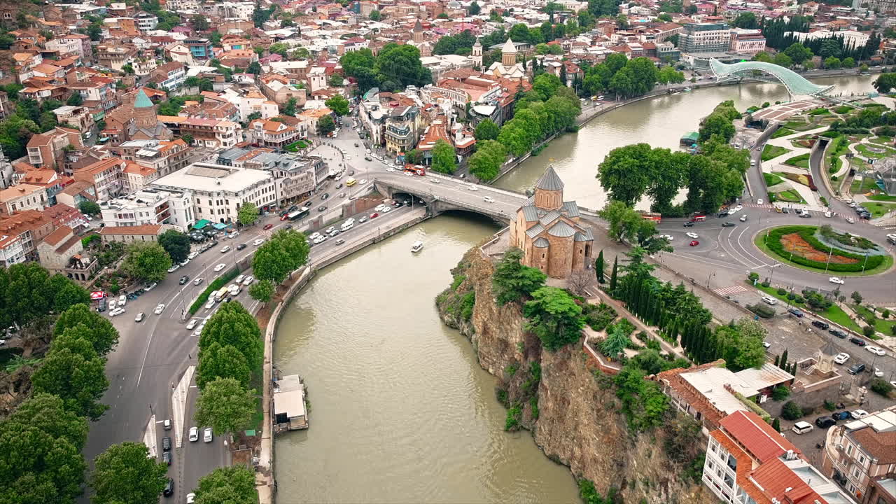 Aerial drone view of Tbilisi, Georgia at cloudy weather. Metekhi Church, water channel, multiple buildings, a lot of greenery, road with traffic