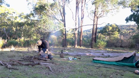 A bushman puts a billy on the boil with his swag set up in the Australian bush