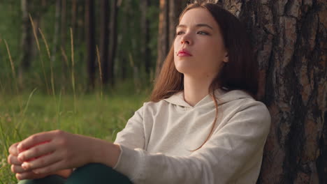 la mujer piensa sentada junto a un árbol en el bosque. los rayos del sol caen sobre la mujer deprimida descansando cerca del tronco de la planta rodeada de naturaleza salvaje. actitud pesimista hacia la vida.