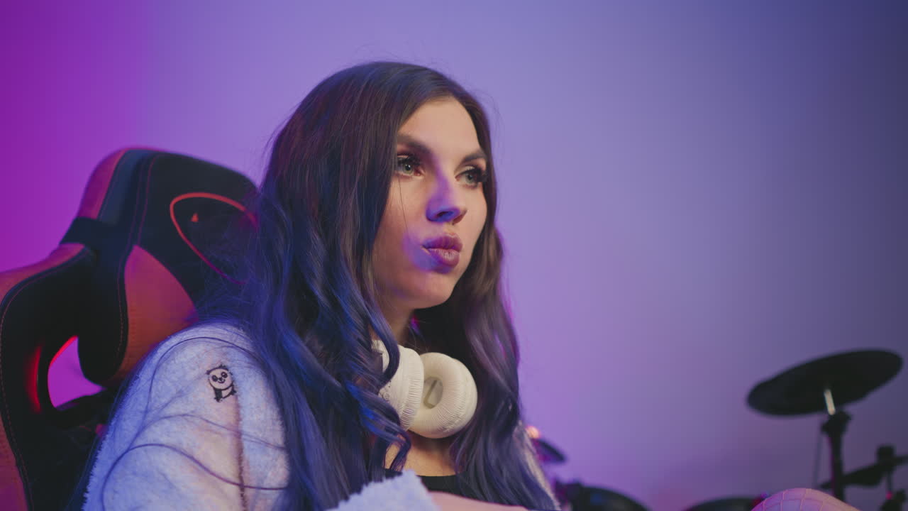 Young lady with painted eyes leans back in gaming chair while eating popcorn, gazing toward something off screen with soft colored lighting and electronic drum kit visible behind her