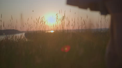 a shot of tall grasses swaying gently at sunset with a lake in the background, as the lower body of a woman passes by, casting a warm, glowing light. the scene exudes tranquility and natural beauty