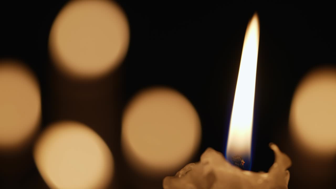 Candela di cera accesa che tremola nel buio su uno sfondo nero con effetto bokeh sfocato della luce della candela. Sfondo o illustrazione di ricordo, lutto o celebrazione. Primo piano di candele accese in chiesa