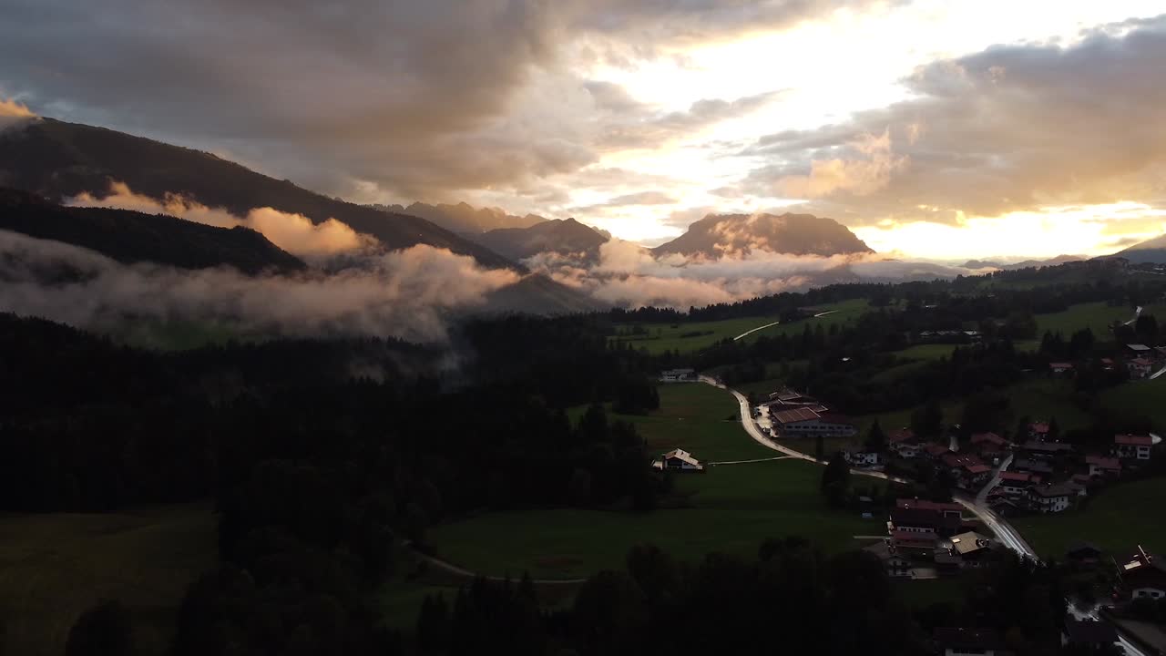 una pintoresca vista de un pintoresco pueblo ubicado en la base de las montañas, con nubes bajas que proyectan sombras durante la puesta de sol