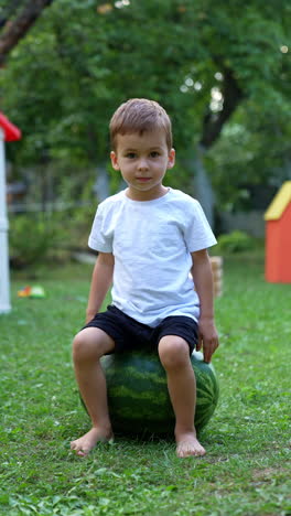 charming caucasian toddler sitting on the big watermelon in the garden. kid claps his hand by the berry. vertical video.