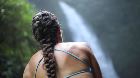 Slow-motion-panning-shot-of-a-girl-in-a-blue-bikini-sitting-in-front-of-a-gushing-NungNung-Waterfall-in-Bali,-Indonesia