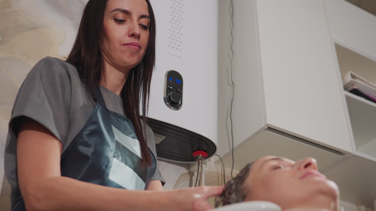 Peluquera blanca enjuagando el pelo en el lavabo del salón, espuma suave y flujo de agua, peluquera morena aplicando limpiador y masajeando, toalla y capa visibles, clienta reclinada disfrutando de un lavado de pelo tipo spa