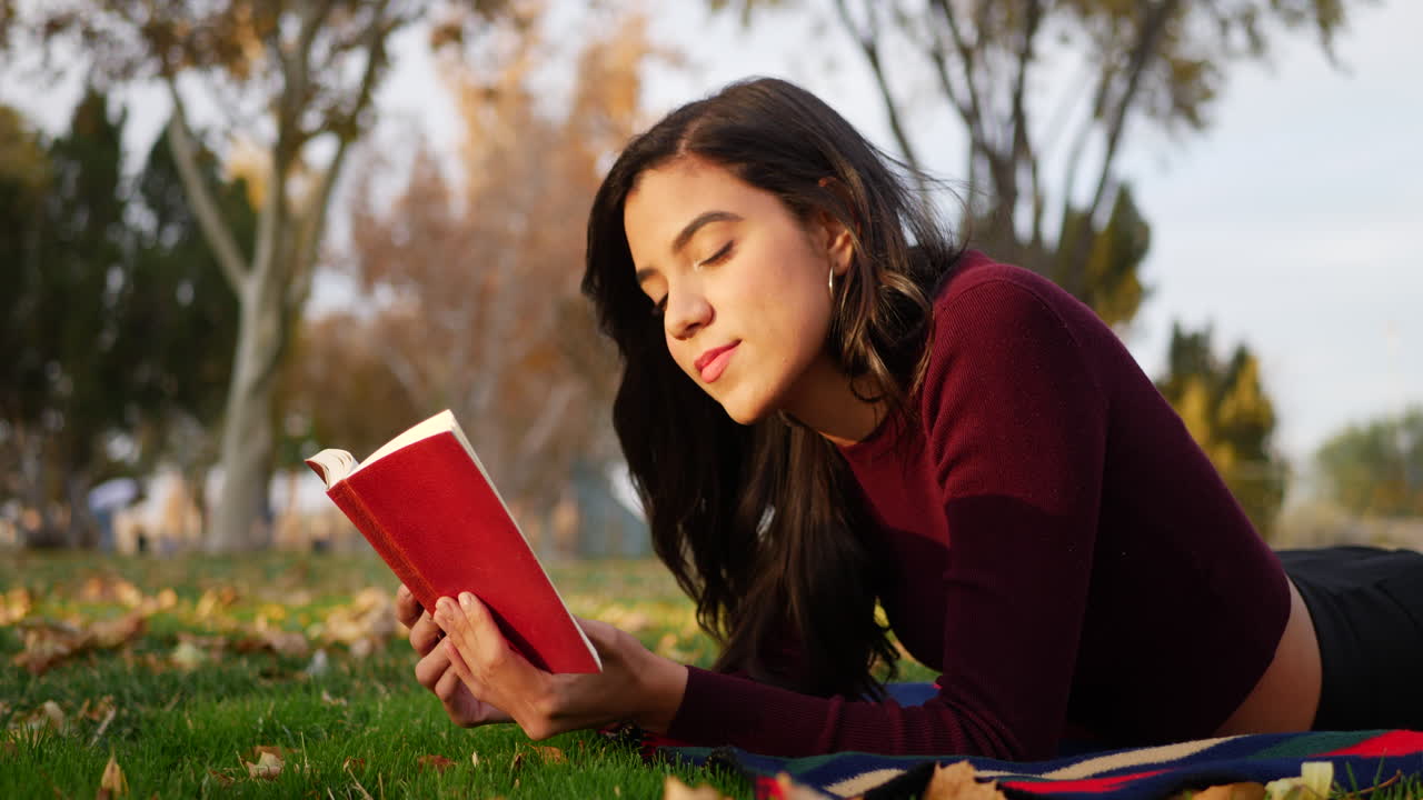una estudiante universitaria hispana leyendo un libro rojo y sonriendo mientras se acostaba en el césped de un parque al atardecer