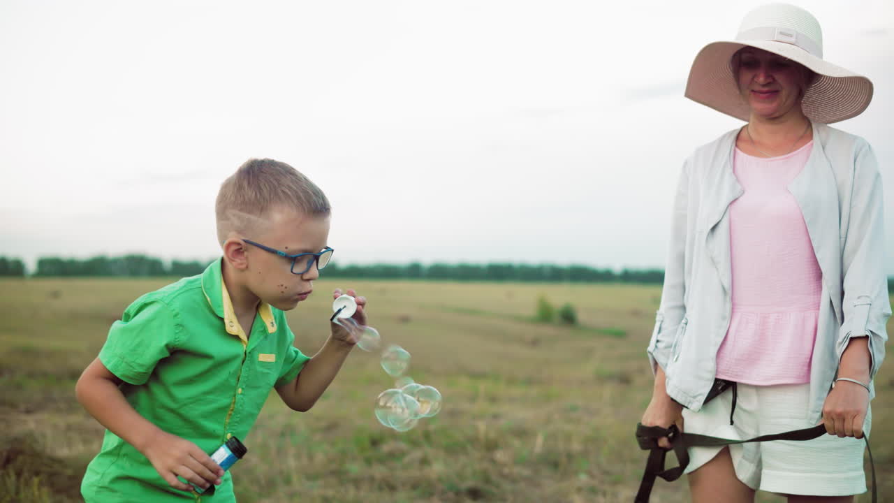 초록색 셔츠를 입은 작은 소년은 조심스럽게 거품을 불어넣고 개 주인이 열렬한 애완동물을 아다니는 것을 막고 있습니다. 광활한 농경지, 어져있는 풀, 따뜻한 자연 조명