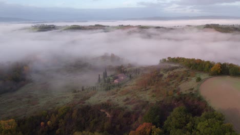 Tuscany-valley-filled-with-mist-in-morning,-small-rural-farm-house,-aerial