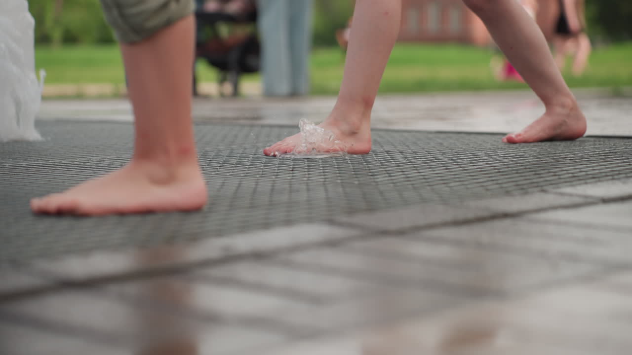 Leg view of barefoot children using legs to close water fountain jet on metal grate, blurred people in background, park setting, summer play turning spray off, curious interaction