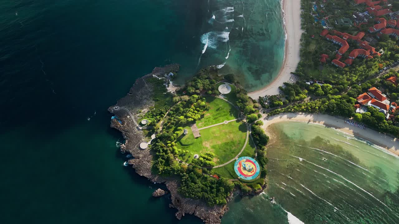 Aerial drone footage of the peninsula at Bali during golden hour showing turquoise lagoon beside a sandy beach with a coastal park and resort area while open ocean surf breaks along the shoreline