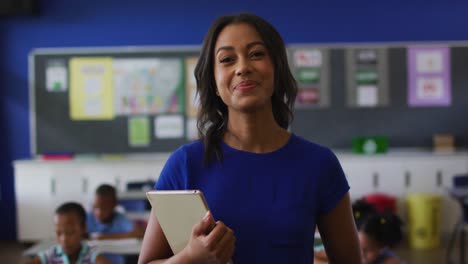 Portrait-of-happy-mixed-race-female-teacher-standing-in-classroom-with-tablet-children-in-background