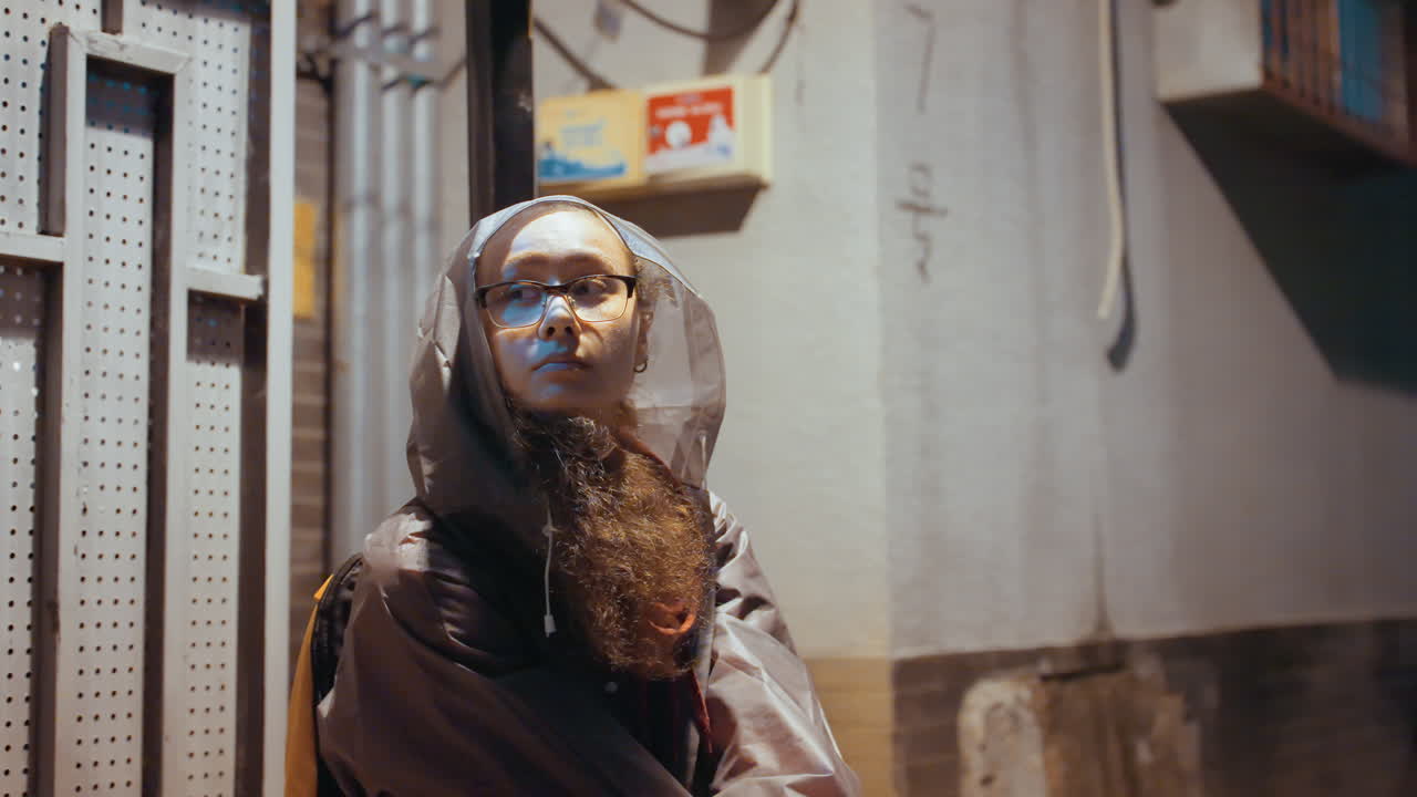 Ritratto notturno: viaggiatore contemplativo con cappuccio e occhiali, barba lunga illuminata dal lampione, seduto contro un cancello di metallo, effetto grana cinematografica vintage, tenue bagliore al neon, ronzio di scooter in lontananza, poeta errante.