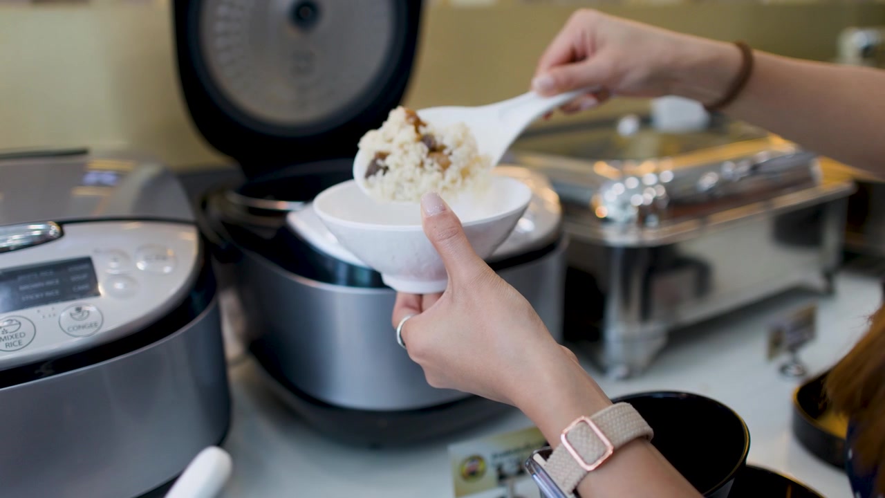 Close-up of hands scooping white rice from a professional cooker into a small ceramic bowl