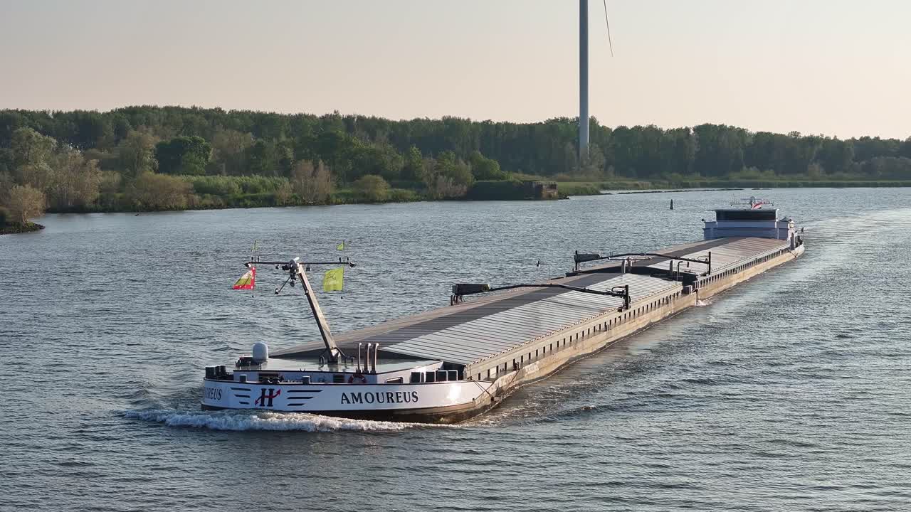 A long cargo ship named 'AMOUREUS' navigating a river with a wind turbine in the background