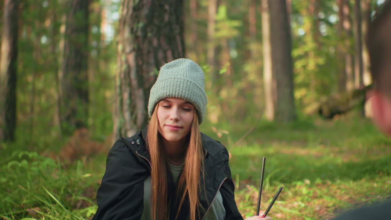 Girl holding tent poles smiles while teasing her brother before handing them over during forest camping activity, both enjoying lighthearted moment in natural woodland setting