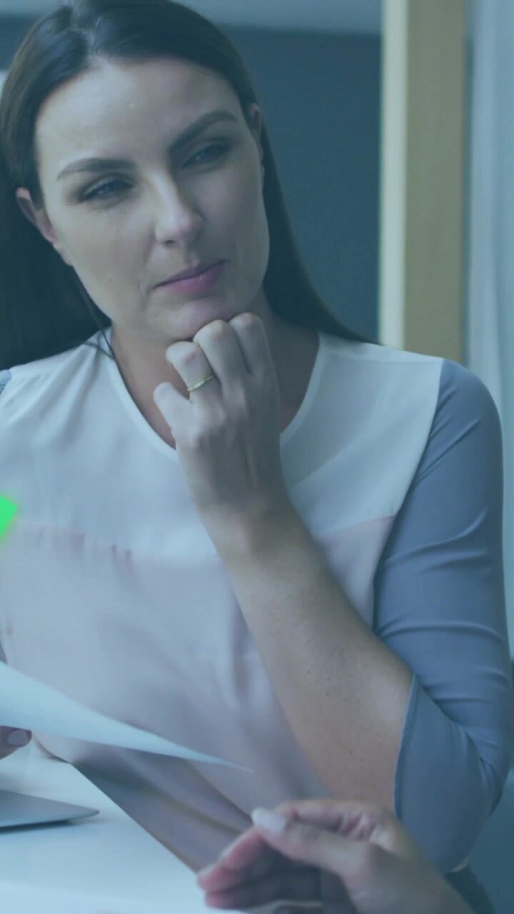 Video verticale: Donna che parla di finanza dopo un cenno di un collega, mostrando un foglio con grafici al neon sovrapposti.
