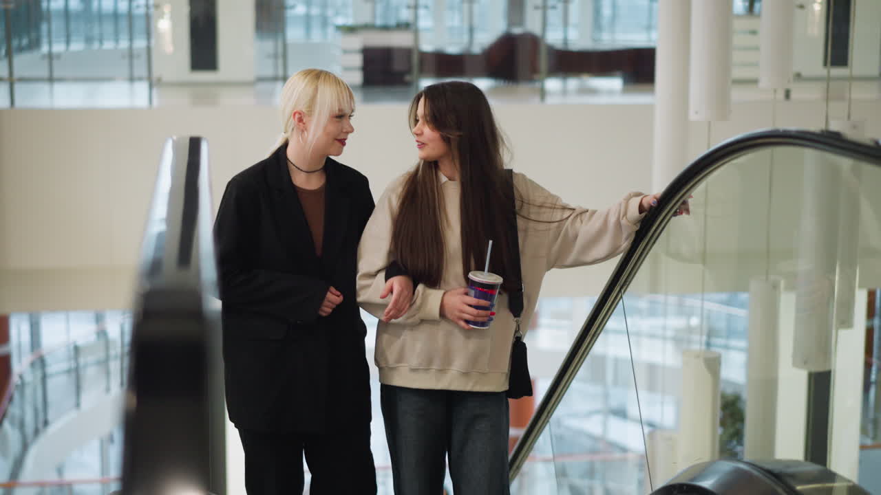 High school students ascend escalator in shopping mall while interacting, smiling, one holding drink, both enjoying lively conversation in bright modern retail environment