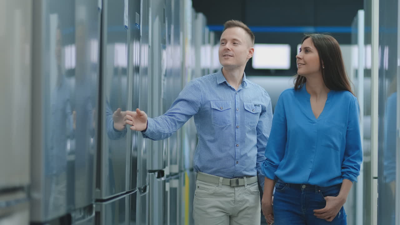 A young couple, a man and a woman choose a new fridge. They open the door and look at the spaciousness and convenience.