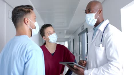 diverse doctors wearing face masks, discussing work and using tablet in corridor, slow motion