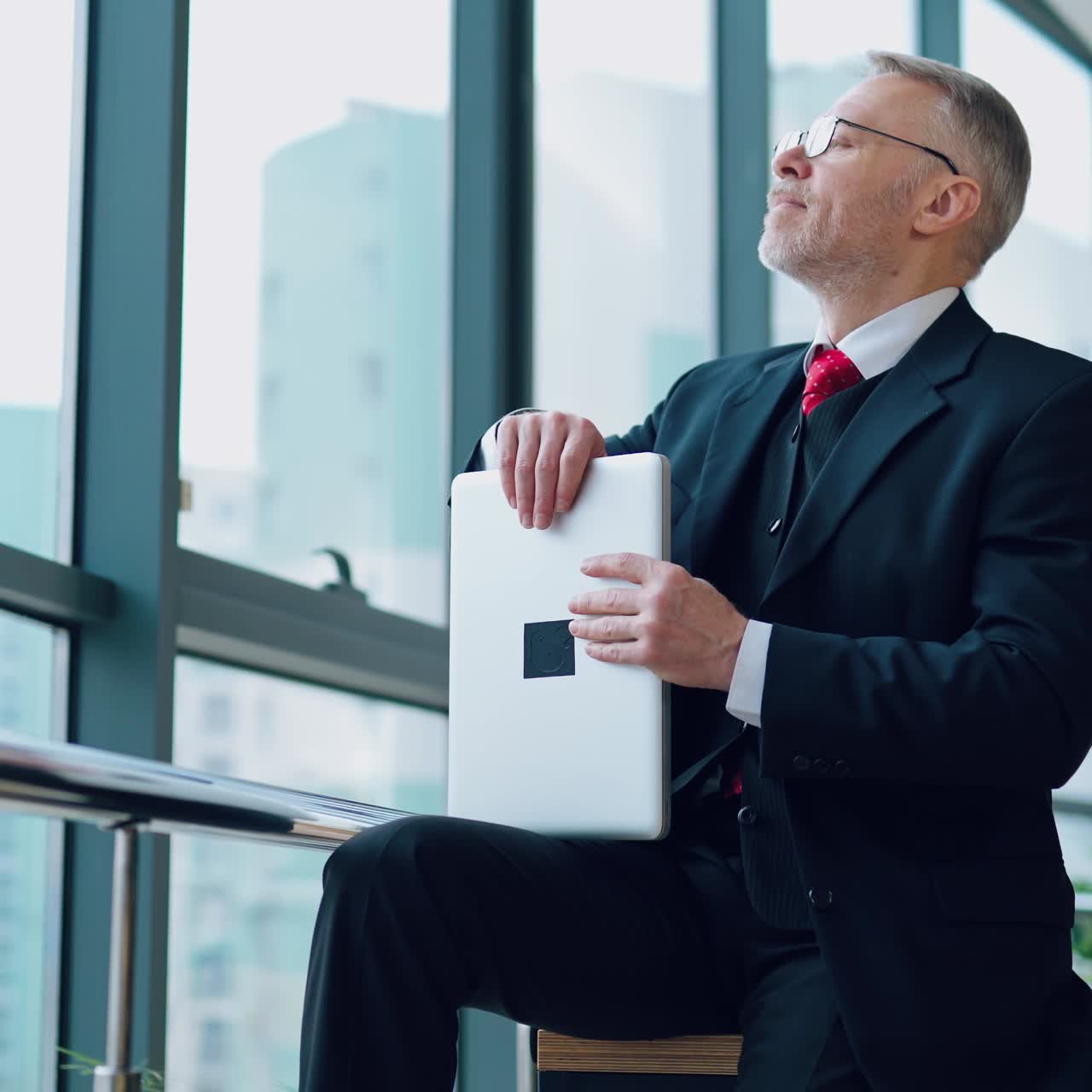 Senior businessman in suit working near office window. Elderly elegant man is typing on a laptop while sitting near the window. Business concept.