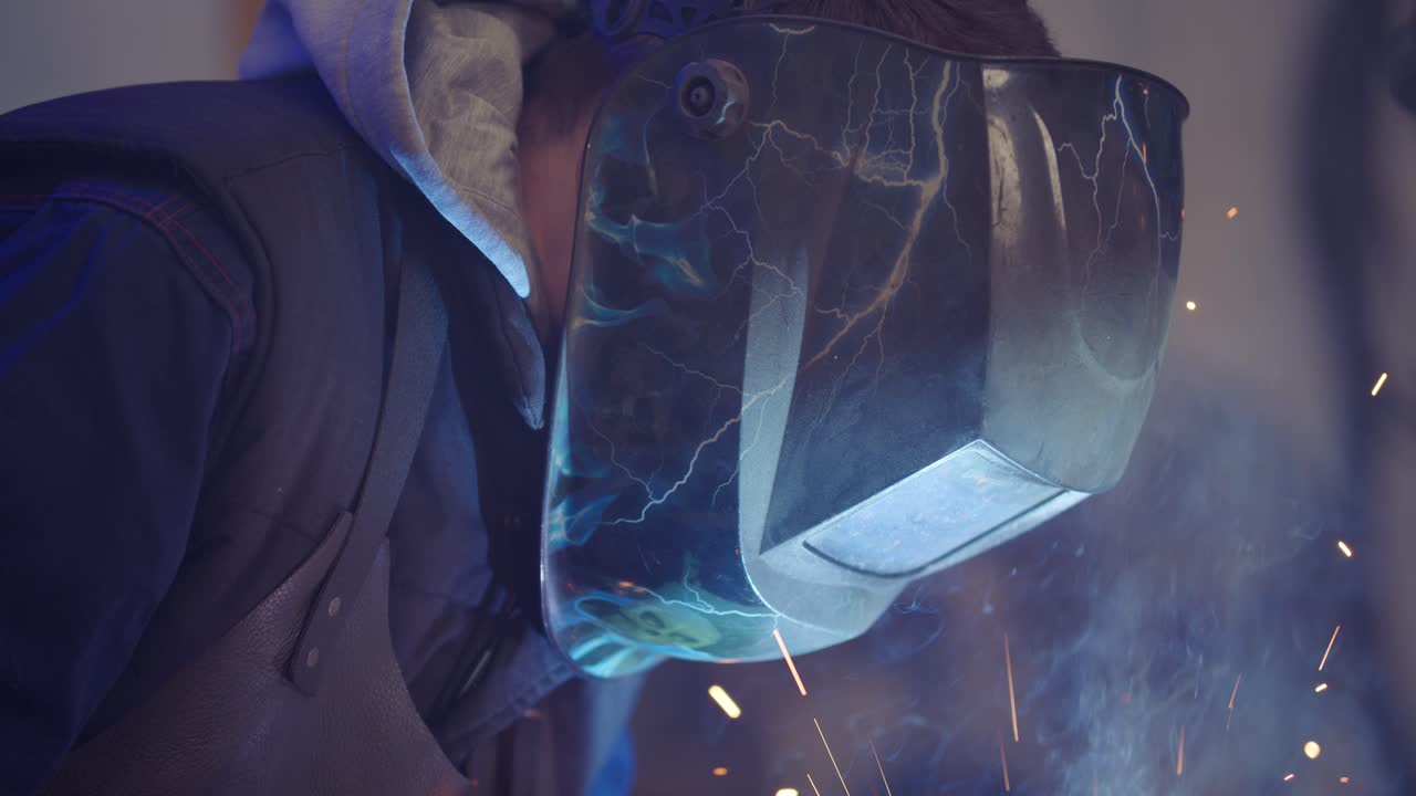 Close up of a helmeted welder welding metal hinges while sparks fly in slow motion