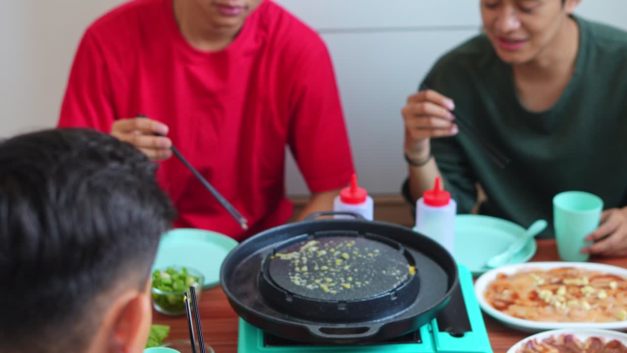 Group Of Young Asian Men Gather Around Table Enjoying Casual Homemade Meal Cooked On Portable Grill