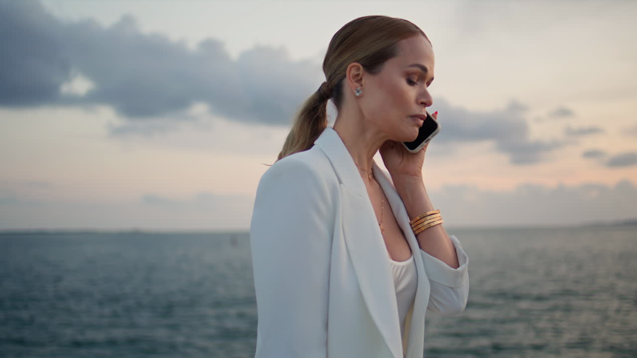 우아한 비즈니스 레이디는 바다의 해가 지는 곳을 가까이에서 걸어다니고 있습니다.