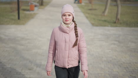woman dressed in pink winter jacket and beanie walks along paved park path on cold sunny day, looking to side, surrounded by sparse leafless trees and soft natural light in peaceful urban setting