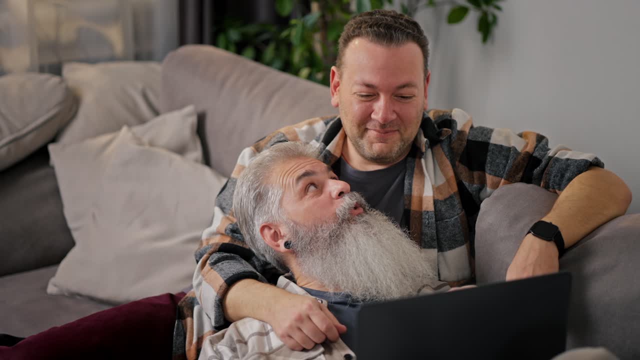 A happy brunette man with stubble in a checkered shirt watches a video using a gray laptop together with his elderly boyfriend with gray hair and a lush beard lying on the sofa in a modern apartment