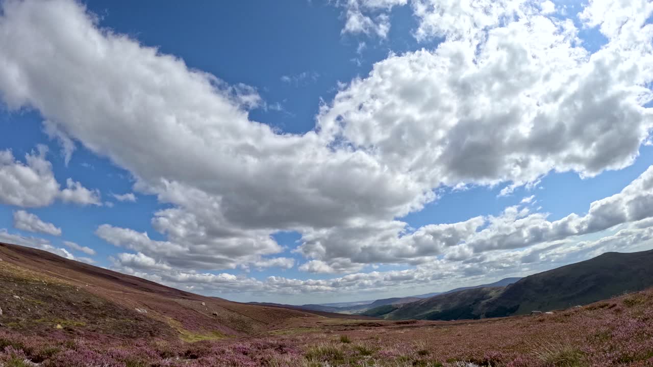 Dynamic clouds drift across a bright blue sky above heather-covered hills in Glen Clova, Scotland, captured in a wide-angle time-lapse sequence