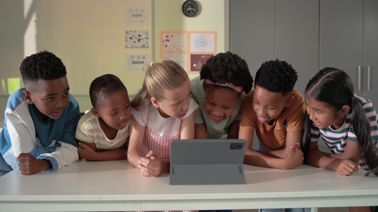 Diverse children leaning toward tablet lighting up, tapping and swiping to share laugh in classroom