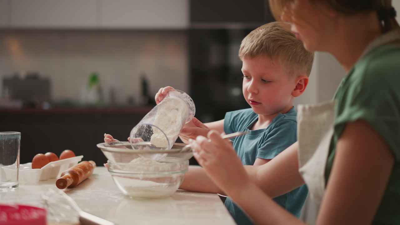 boy holds transparent jar of flour, pouring it into sieve held by woman while looking up and speaking to her, surrounded by baking ingredients and utensils on kitchen counter