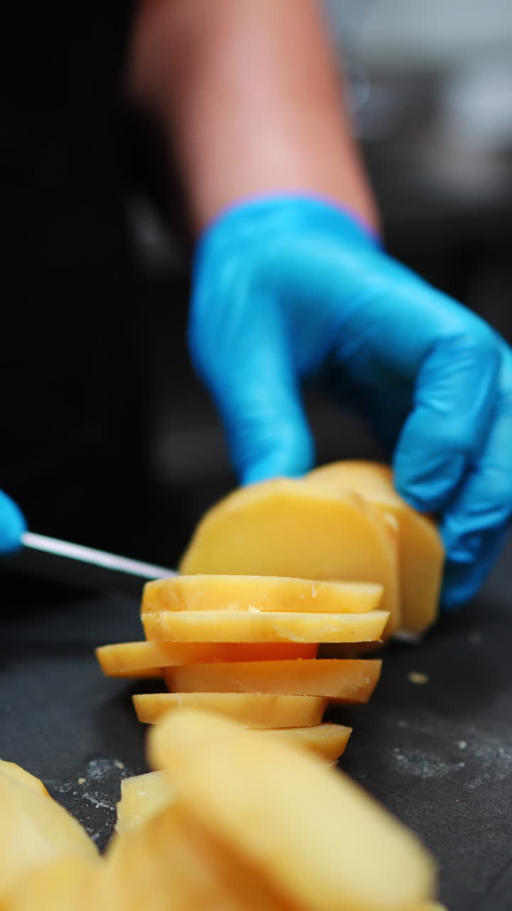 Professional chef wearing blue gloves carefully slicing potato into uniform thin pieces on black cutting board, showcasing precise culinary technique and professional food preparation skills