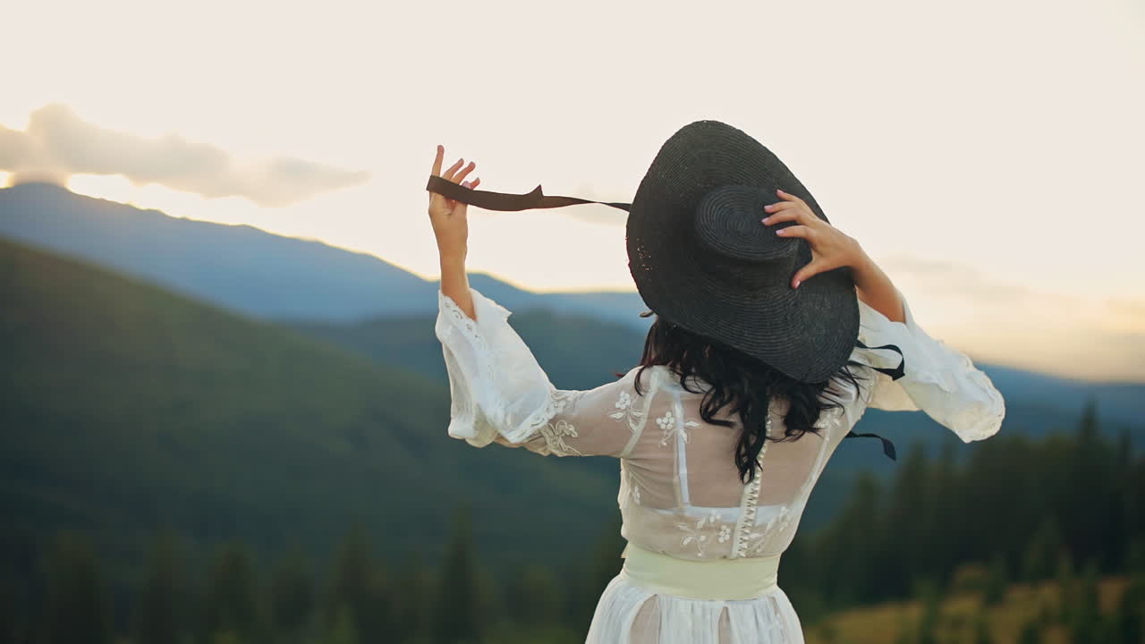 Young lady in white dress and black hat stands her back to the camera facing setting sun. Woman holding her hat string fluttering in the wind. Mountains in blur at backdrop.