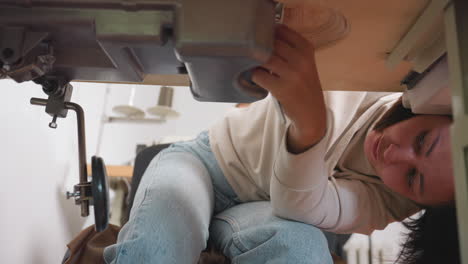 focused tailor bends under sewing machine adjusting internal mechanism from bottom view, showing precision, dedication, and mechanical work in tailoring workspace with fabric equipment