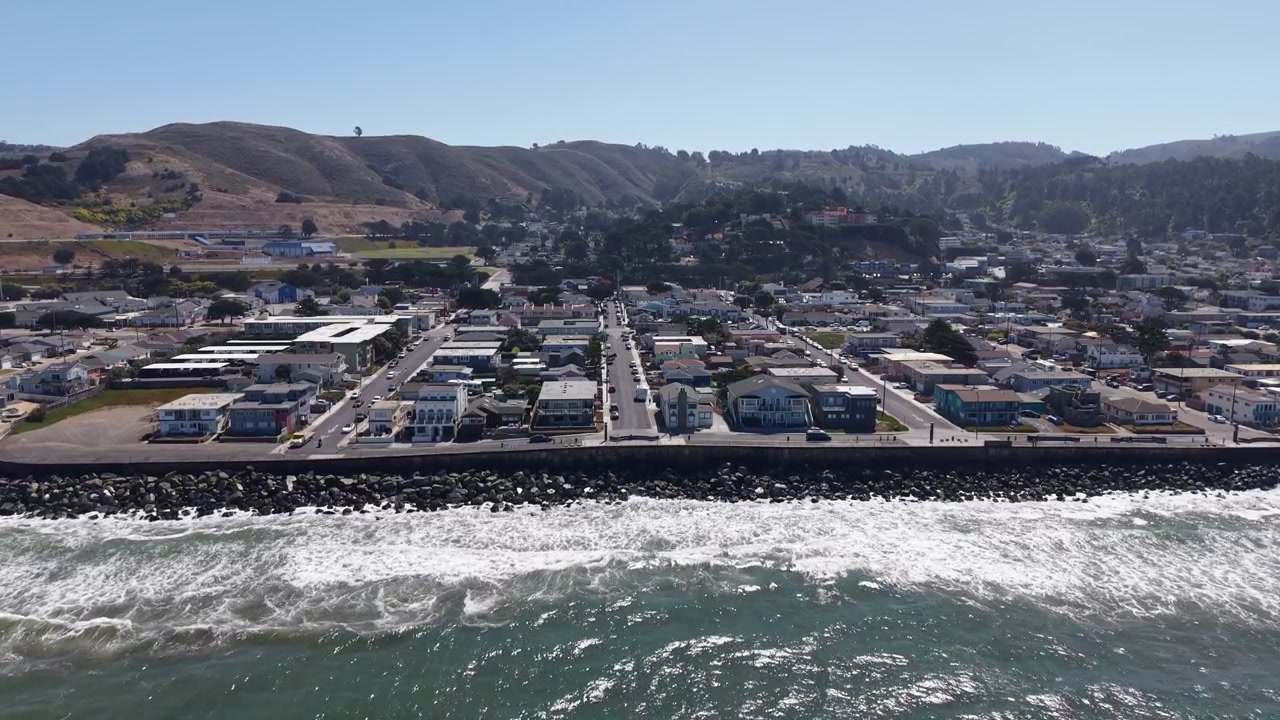 Perspectiva aérea sobre el encantador pueblo costero de Pacifica, California, mostrando la hermosa costa donde las olas rompen contra una barrera de rocas artificial. Toma de dron descendente