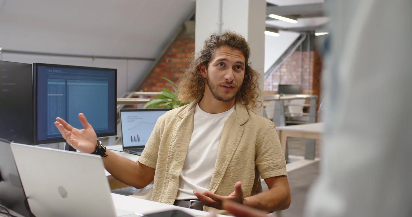 Adult man listening and responding to standing coworker in loft office, pointing at laptop charts