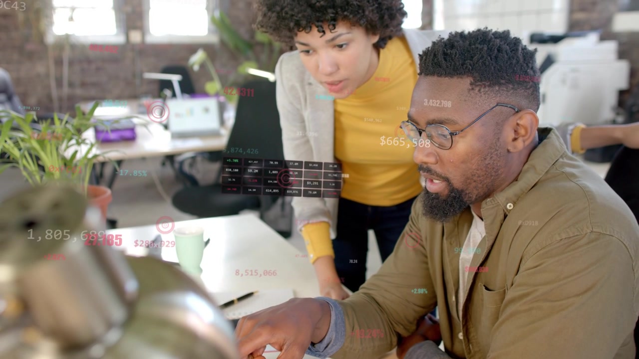 Female leaning, pointing at laptop, leading male tapping trackpad, discussing business with labels