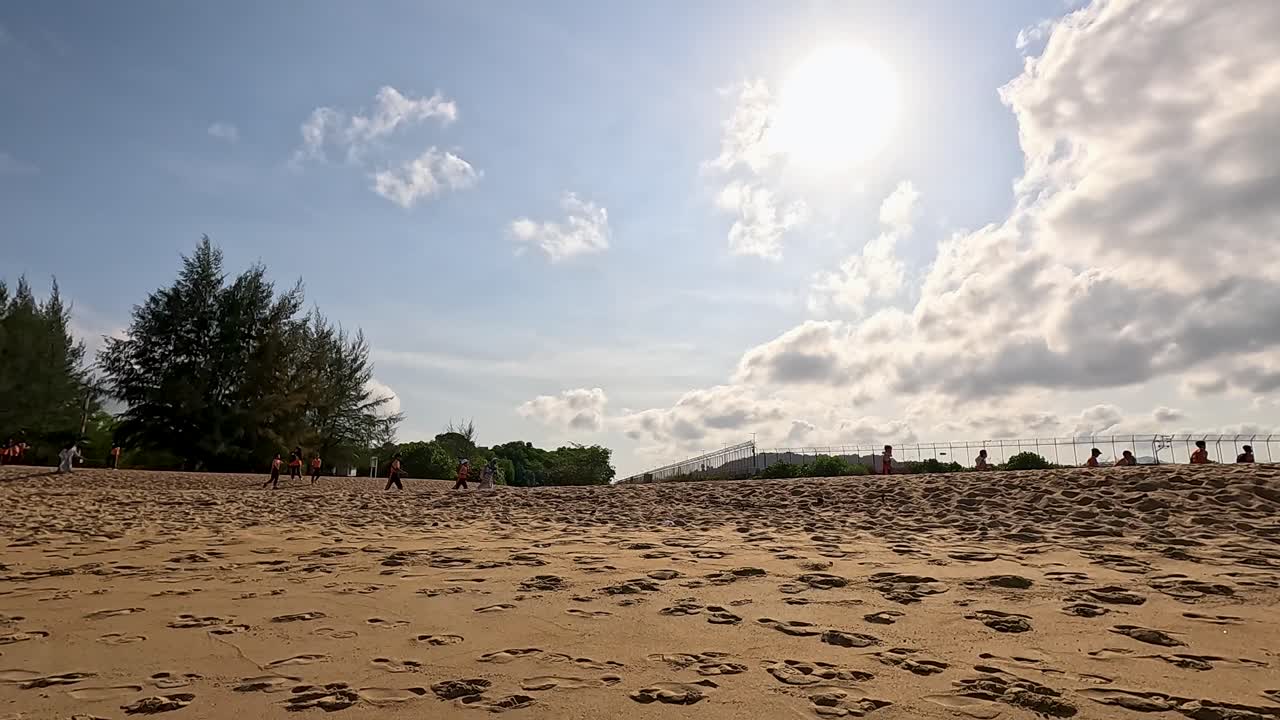 Children walk along a sandy beach in Phuket under bright sunlight, with trees and clouds in the background