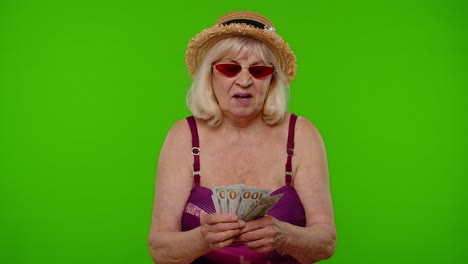 a senior woman wearing a hat and sunglasses smiles while holding us dollar bills