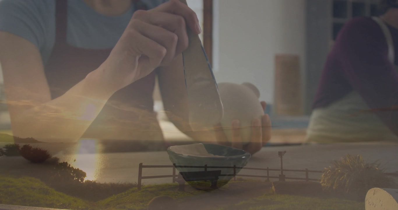 Pottery colleagues smoothing ceramic vase with metal tool, showing transparent sunset lake overlay