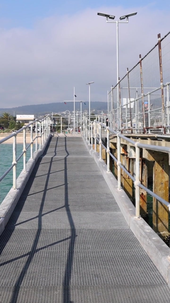 First-person view walking along a narrow concrete pier with metal railings under blue sky.
