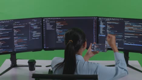 back view of happy asian woman developer looking at code on mobile phone while write code with multiple computer screens in green screen studio