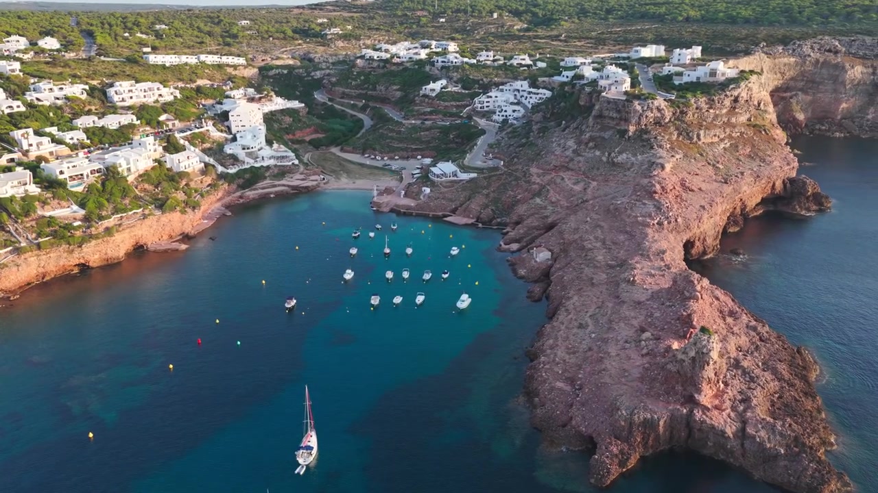 Luftfoto panorering af Cala Morell, Menorca, kystlandsby, bugt, både, middelhavslandskab, strand