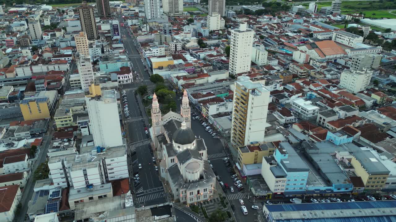 Looking down the main street in Pouso Alegre, Minas Gerais, showcasing the historic cathedral nestled between tall, modern high-rise buildings and dense urban commercial and residential blocks