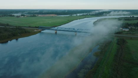 Aerial-establishing-view-of-the-steel-bridge-over-Lielupe-river-on-a-sunny-summer-morning,-fog-rising-over-the-river,-cars-driving,-wide-drone-shot-moving-forward